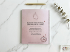 Pink notebook with motivational quote on a marble surface with a pen and plant.